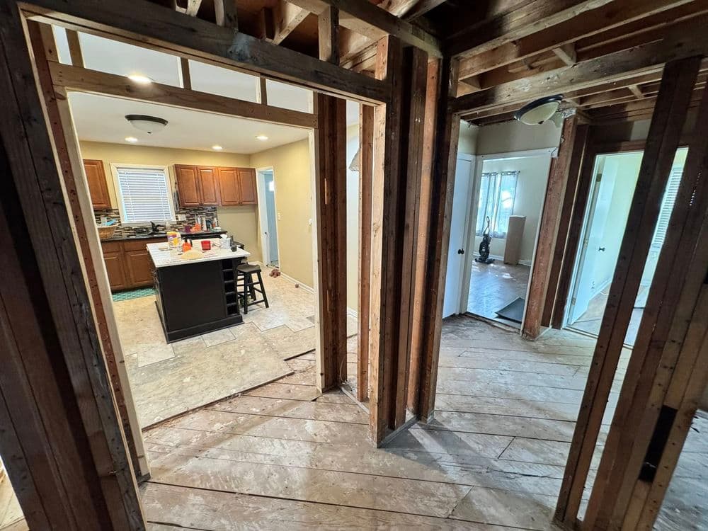 Interior of a home renovation showing exposed wooden framing and open doorways to a kitchen.