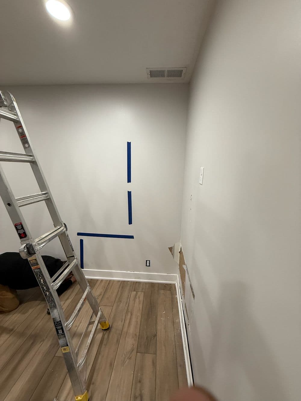 Room renovation in progress with a ladder, blue painter's tape on white wall, and wooden floor.