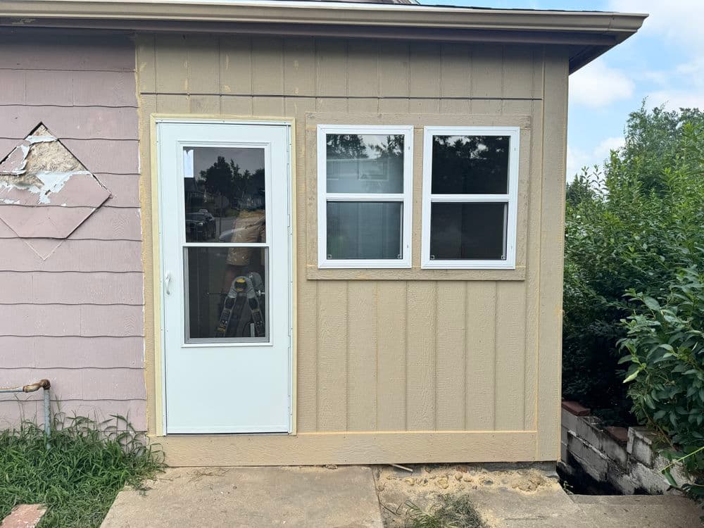 Exterior view of a house showing a new white door and two windows on a tan wall.